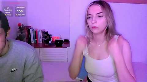 Video of a blonde woman in a white tank top, sitting on a bed with a bookshelf behind her, featuring various books and a vase of flowers.
