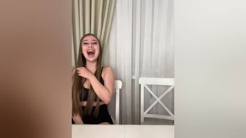 Video of a young woman with long, straight brown hair, wearing a black lace bra, sitting on a white chair, laughing with her hand on her head, in a simple, brightly lit room with white curtains and a white table.