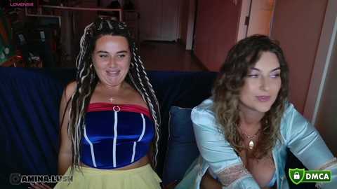 Video of two women, one in a blue strapless top with yellow skirt, the other in a light blue top, sitting on a dark blue couch.