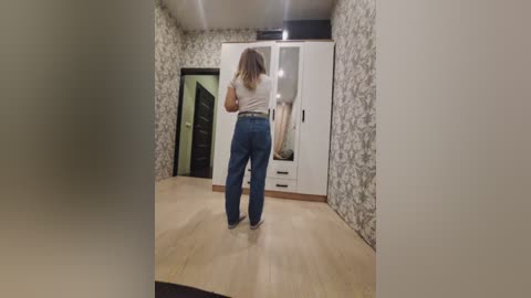 Video of a woman with shoulder-length blonde hair in a white top and blue jeans standing in a modern, minimalist bedroom with light wood flooring, white patterned wallpaper, and a large white wardrobe.