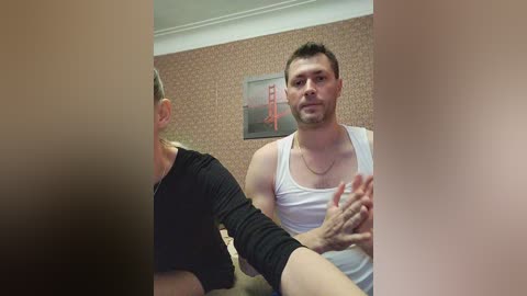 Video of a muscular, light-skinned man in a white tank top, speaking to a partially visible woman in a black top, in a beige-patterned room.