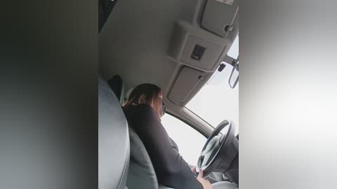 Video of a woman with shoulder-length brown hair, wearing a black top, sitting in a car with a grey dashboard, steering wheel, and beige interior.