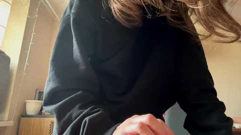 A video of a person in a dark green sweater, bending over, with long, wavy brown hair partially covering their face. The background shows a room with a beige wall, a wooden cabinet, and a string of lights.