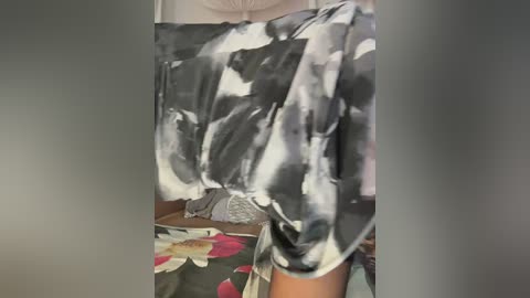 A video of a person wearing a patterned, sleeveless top with a monochromatic, abstract floral design. The background features a couch with a floral-patterned cushion and a partially visible, blurred person.