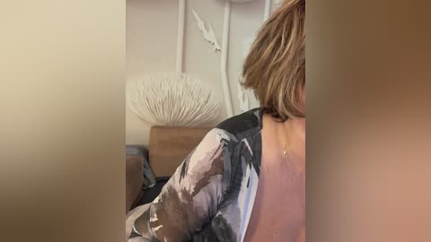 Video of a partially visible person with shoulder-length brown hair, wearing a black and white patterned shirt, sitting on a brown couch in a room with a white seashell decoration and white wall art.