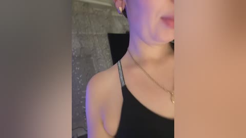 Video of a woman with fair skin and short hair, wearing a black spaghetti strap top, and a gold necklace, in a dimly lit room with a textured grey wall.