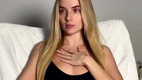 Video of a young woman with long, straight blonde hair, fair skin, and green eyes, wearing a black tank top, seated on a white, textured cushion, with a neutral background.