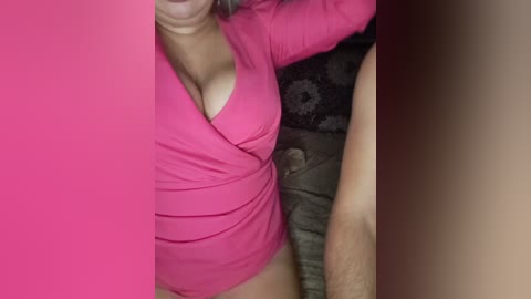 Video of a woman in a tight, bright pink, long-sleeve bodysuit with a plunging neckline, revealing ample cleavage. Her fair skin and shoulder-length hair are partially visible. Background includes a dark, patterned cushion and a light-colored arm.