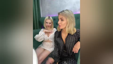 Video of two women seated on a green velvet couch. The blonde woman in a white lace dress and the brunette in a black pinstripe dress look at each other.