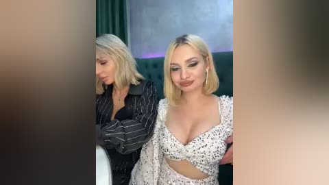 Video of two blonde women seated indoors; one wears a black pinstripe blazer, the other a white polka-dot crop top, both smiling.