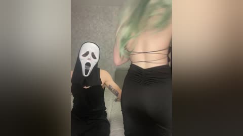 Video of a person wearing a white mask with a black hood, black pants, and a black tank top, standing beside a person with green hair in a black strappy top, in a dimly lit room.