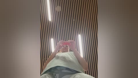 Video of a young woman in a hospital gown, hands clasped, looking upward through a narrow slit in the ceiling. The background features horizontal wooden slats and fluorescent lights.