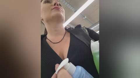 Video of a woman with light skin and dark hair, wearing a black V-neck top and black necklace, holding a white bottle, in a bright, industrial setting with fluorescent lighting.