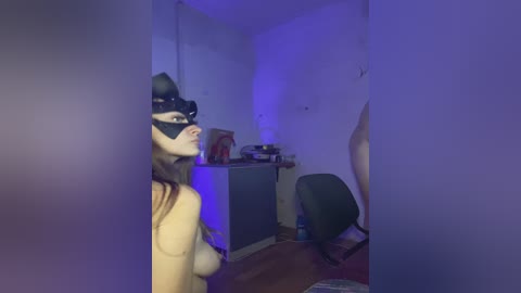 A video shows a person in a black mask and robe, standing in a dimly lit room with blue lighting. The room features a desk, chair, and cardboard boxes.