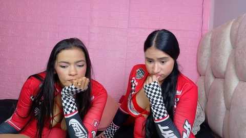 Video of two Latina women in matching red racing suits with black and white checkered gloves, sitting side by side on a black couch against a pink brick wall.