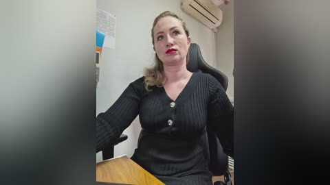 Video of a middle-aged Caucasian woman with light skin and shoulder-length blonde hair, wearing a black ribbed sweater and sitting in a black office chair, looking contemplative.
