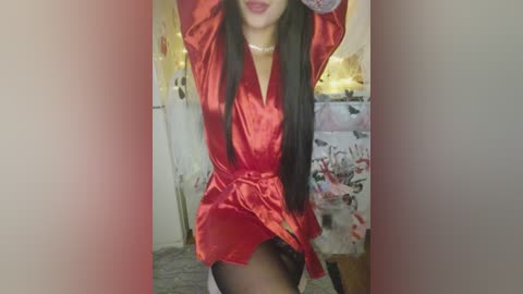 Video of a woman with long black hair, wearing a red satin robe, arms raised, revealing black stockings. Background shows a room with a white door, a dresser with floral pattern, and a bed with a white sheet.