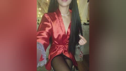 Video of an Asian woman with long black hair, wearing a red satin robe with lace sleeves, a pearl necklace, and black stockings, sitting on a bed with fairy lights in the background.