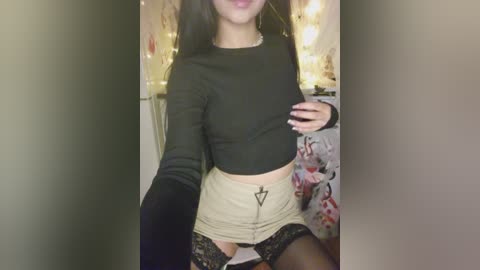 Video of a woman with long dark hair, wearing a black crop top, beige mini skirt with lace, and black thigh-high stockings, standing in a cozy room with fairy lights and floral-patterned bedding.