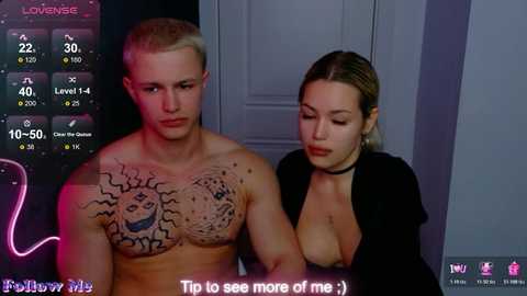 Video: A shirtless, muscular, tattooed man with short blond hair stands next to a blonde woman in a black choker. They are in a dimly lit room with a purple neon sign.