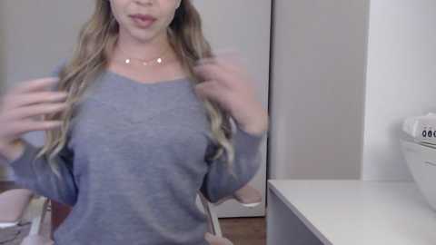 Video of a young woman with long, wavy blonde hair, wearing a grey sweater, posing in a room with a white counter and a beige wall.