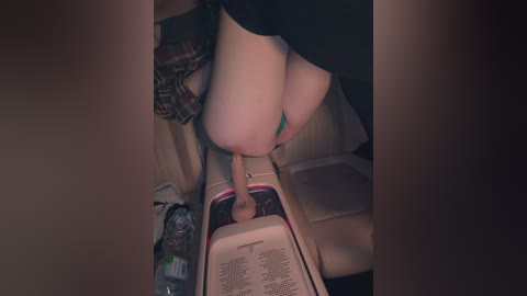 A video shows a person with fair skin wearing a green thong, squatting over a white toilet with a red and white toilet seat. A plaid shirt hangs on the toilet's backrest. A water bottle sits on the floor.