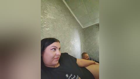 Video of a middle-aged woman with fair skin and dark hair, wearing a black T-shirt, sitting in a dimly lit, concrete room. A man with short hair and dark skin is lying beside her, wearing a white shirt.