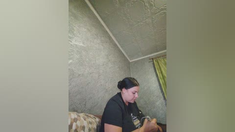 Video of a middle-aged woman with fair skin and dark hair tied back, wearing a black t-shirt, sitting on a patterned sofa in a room with textured gray walls and a green curtain.