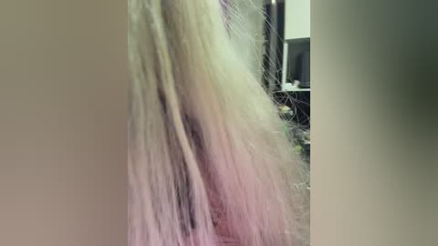 A close-up video of a person's long, light-colored hair with pink and white streaks, viewed from the side, showing a textured, wavy appearance. The background is blurred, featuring an indistinct indoor setting.