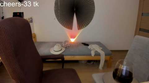 Video of a cluttered living room with a large, decorative black and red fan in the background, a chair, and a table with a hat, clothes, and a wine glass.