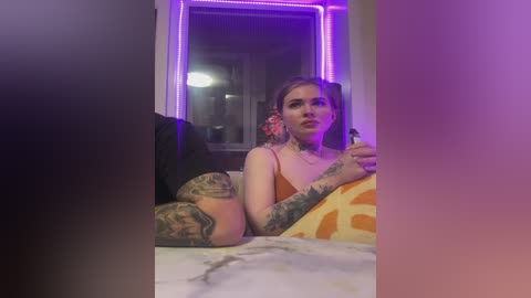 Video of a tattooed woman with long brown hair, wearing a sleeveless orange top, reclining on a bed. Neon purple lights illuminate the room, creating a modern, intimate ambiance.