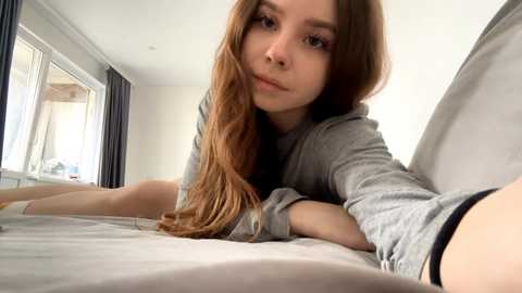 A young, fair-skinned woman with long, wavy brown hair and light makeup, wearing a gray hoodie, lies on a bed, gazing directly at the camera. The room has white walls, a window, and a gray blanket.