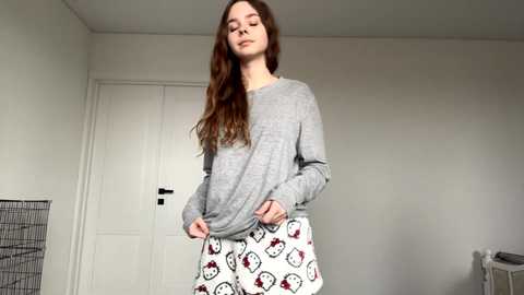 Video of a young, fair-skinned woman with long brown hair wearing a grey sweater and white pajama pants with red hearts and cartoon characters, standing in a simple, light-colored room with a white door and a metal crib in the background.