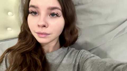 Video of a young woman with long, wavy brown hair and fair skin, wearing gray clothing, lying on a light-colored, tufted headboard. She has long eyelashes and a neutral expression.