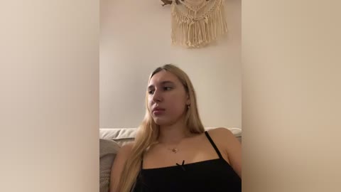 Video of a young woman with long blonde hair, wearing a black spaghetti-strap top, sitting on a beige couch in a dimly lit room with a macram\u00e9 wall hanging.