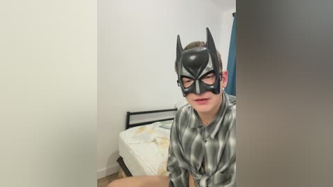 Video of a young man in a Batman mask and gray, checkered shirt, sitting on a bed in a simple, white-walled room with a blue curtain.