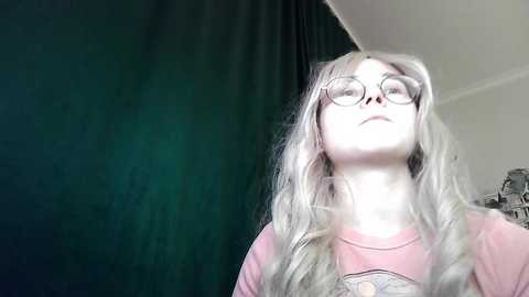 A video of a young woman with long, wavy blonde hair, wearing glasses and a pink graphic tee. She stands against a dark green curtain with a white wall in the background.