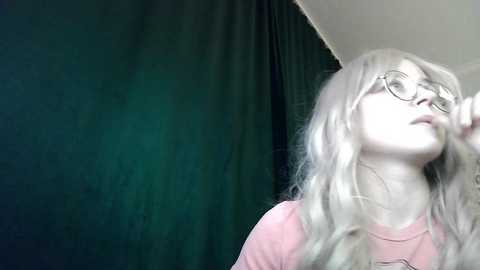 Video of a young woman with long, wavy, platinum blonde hair and round glasses, wearing a pink t-shirt. She has a thoughtful expression, looking upward. The background features a dark green curtain and a white ceiling.