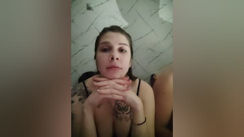 Video of a young woman with light skin, brown hair tied back, wearing a black bra, sitting on a bed with a white marble-patterned wall in the background.