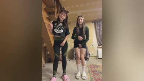 Video of two young women in a bedroom. One in a black crop top and leggings, the other in a long-sleeve top and shorts. Background shows a messy room with exposed pipes and a patterned rug.