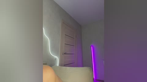 Video of a dimly lit room with a beige sofa, wooden door, and a glowing neon light on the wall. A purple neon stick stands in the background, casting a soft glow.