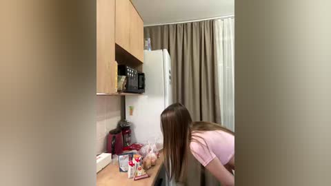 Video of a small, cozy kitchen with beige walls and wooden cabinets. A young girl with long brown hair, wearing a pink shirt, leans over a countertop, possibly cooking.