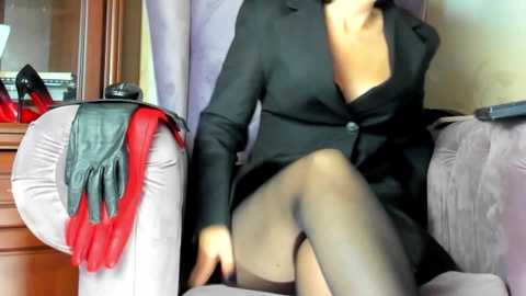 Video of a woman in a black blazer, sheer black stockings, and a red scarf, sitting on a gray sofa, next to a wooden dresser.