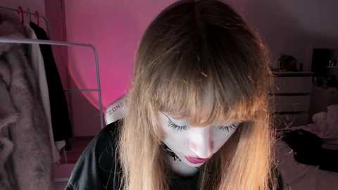 A video of a young woman with long, straight, light brown hair and pale skin, wearing dark makeup and a black choker, sitting in a dimly lit room with pink lighting.