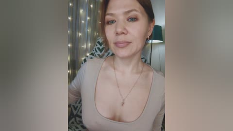 Video of a fair-skinned, middle-aged woman with light brown hair and blue eyes, wearing a beige top that accentuates her medium-sized breasts. She's smiling, surrounded by a cozy, dimly lit room with string lights and a patterned blanket in the background.