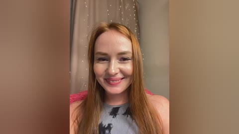 Video of a young Caucasian woman with long, straight red hair, wearing a tie-dye top, smiling, standing in front of a beige curtain with small white dots.