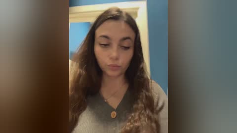 Video of a young woman with long brown hair and fair skin, wearing a green top, standing in a room with blue walls and a white door.