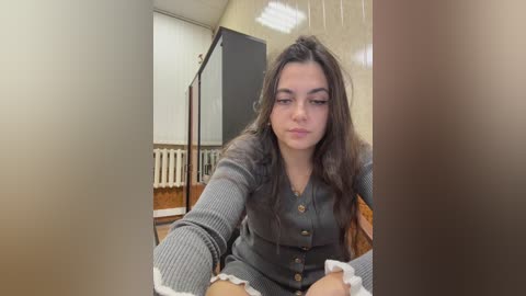 Video of a young woman with long dark hair, wearing a gray ribbed cardigan, seated in a room with beige walls, a radiator, and a window with blinds.