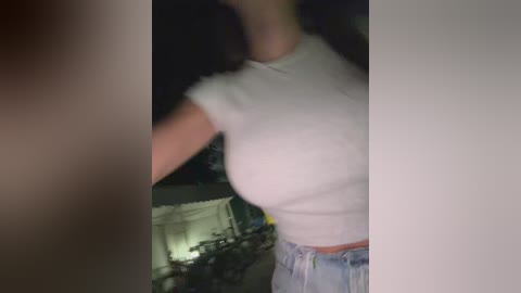 A blurry, dimly-lit video of a woman wearing a tight white T-shirt and light blue jeans, standing indoors with a blurred background.