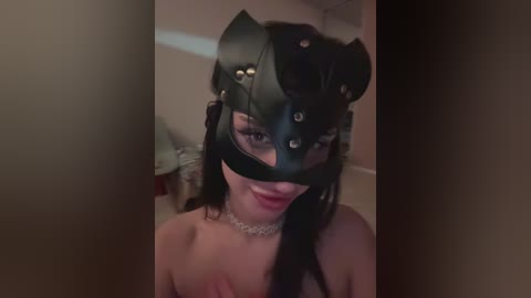 Video of a woman with long black hair, wearing a black cat mask with silver studs and a silver choker, indoors with dim lighting.